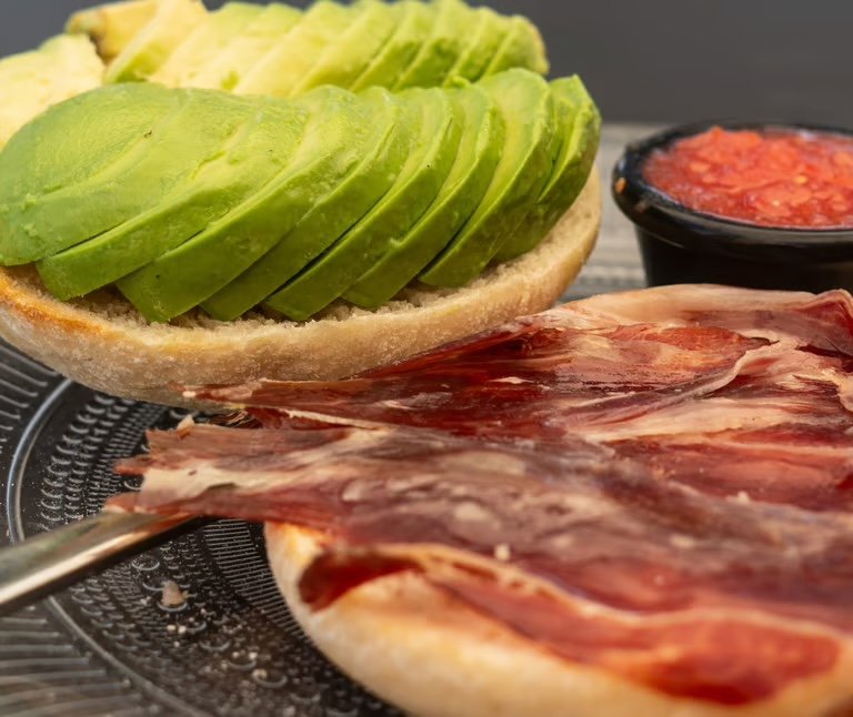 Detalle de tostadas con aguacate, jamón y un cuenco de tomate en Bahía